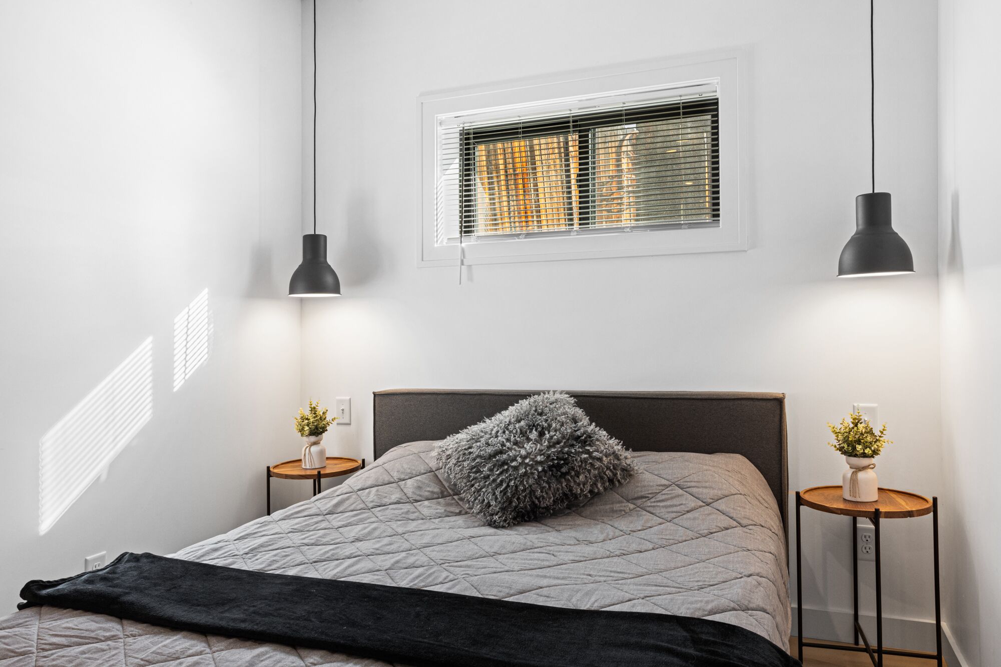 A bedroom with a bed, two bedside tables, a window with blinds, and two pendant lights.