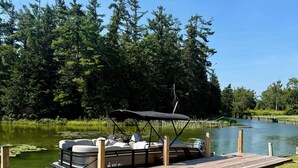 A dock with a pontoon boat, a grassy area, and a lake surrounded by trees.