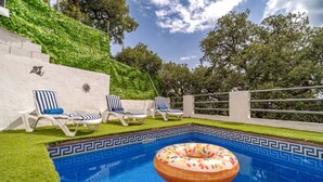 A swimming pool with a donut float, surrounded by white lounge chairs with blue cushions, and a white wall with a decorative sun motif.
