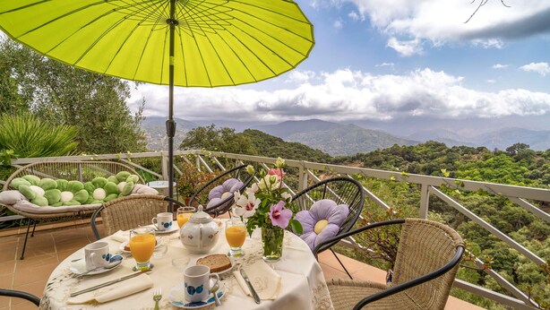 A table set for breakfast with a yellow umbrella, wicker chairs, and a scenic mountain view.