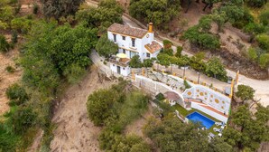 An aerial view of a white building with a tiled roof, surrounded by greenery and a swimming pool.