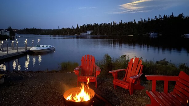 A lakeside setting with a fire pit, red Adirondack chairs, and a dock with a boat.