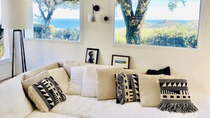 A living room with a beige sofa, multiple pillows, and two large windows with a view of trees and hills. - Luxurious Mas with Bassin Balnéo Sea View in Collioure 12 pers (Collioure)