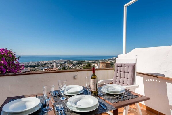 A dining table set with plates, glasses, and cutlery on a balcony overlooking a coastal city. - SHR013 Bonito apartamento con vistas y piscina (Nerja)