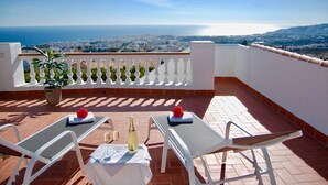 Una terraza con dos sillones blancos, una mesa con una botella de champán y copas, y vistas al mar y al paisaje urbano.