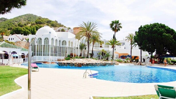 Una piscina con un deck perimetral, palmeras y un edificio con ventanas arqueadas.