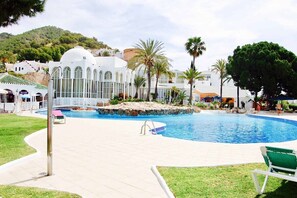 A swimming pool with a surrounding deck, palm trees, and a building with arched windows.