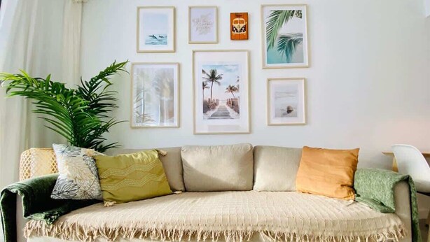 Un sofá color beige con flecos, una planta verde grande y una silla blanca en una habitación con pared blanca y cuadros enmarcados.