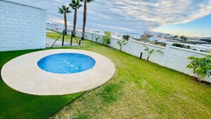 Una piscina de diseño circular, rodeada por un muro blanco y una zona verde con palmeras.