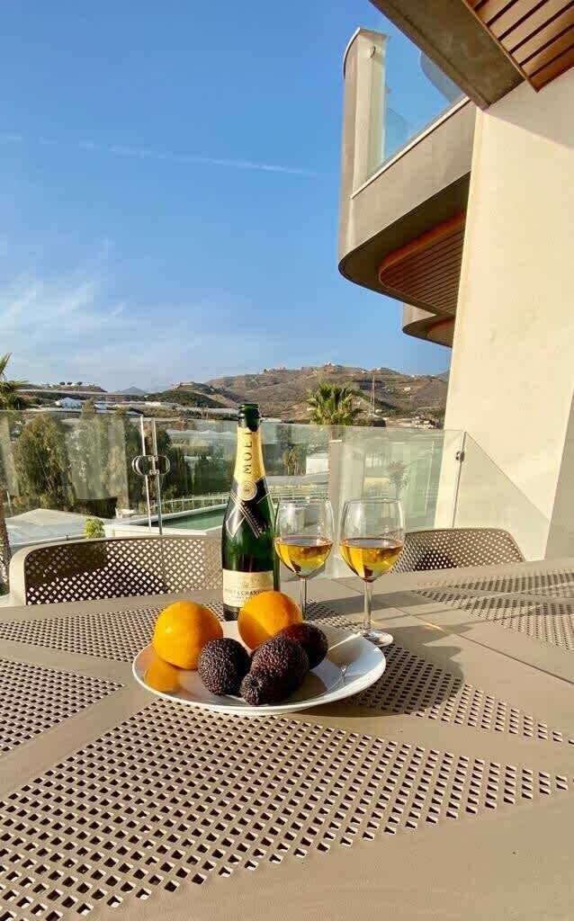 Et bord med en champagneflaske, to glas og en tallerken med frugt på en balkon med udsigt over et landskab.
