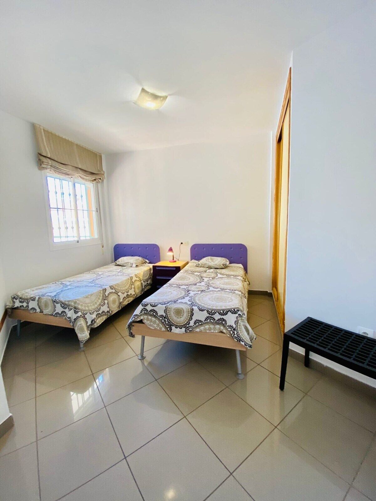 Una habitación pequeña y limpia con dos camas individuales, una ventana con cortinas, una mesita de noche con lámpara y un banco.