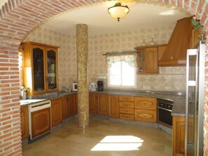 A kitchen with wooden cabinets, a central column, and a window with curtains. - SHR018 Villa El Castillo (Torrox)