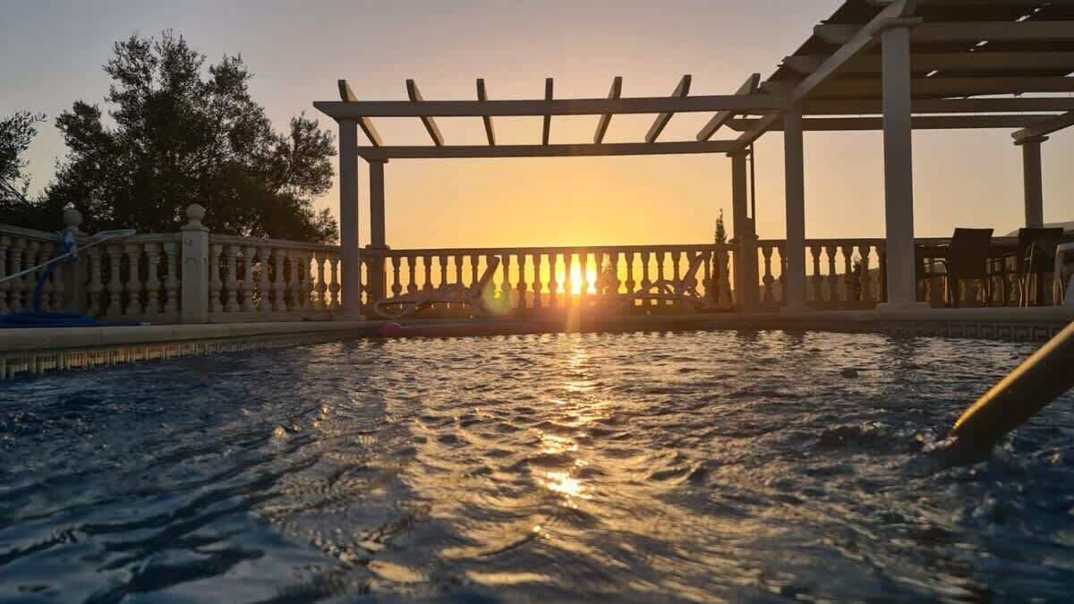 Et poolområde med et pergola-konstruktion, omgivet af et hegn, og en solnedgang i baggrunden.