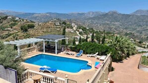 A pool with lounge chairs, a pergola, and a mountainous backdrop.