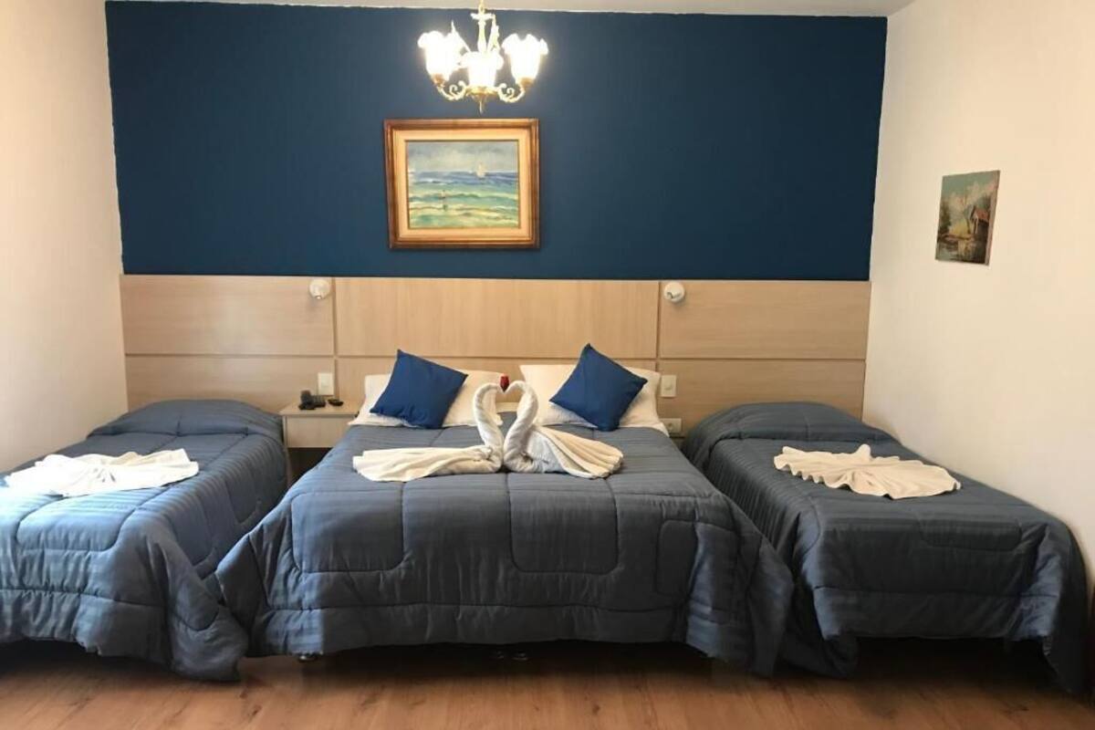Habitación de hotel con tres camas dispuestas en fila, un candelabro y dos cuadros en la pared.