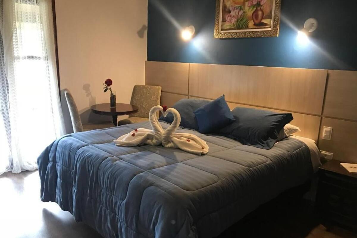 Una cama bien tendida con una colcha azul y almohadas, un arreglo de toallas con forma de corazón, una mesita con un jarrón de flores y un cuadro en la pared.
