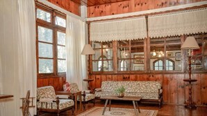 A cozy room with wooden walls, a patterned sofa, a wooden coffee table, and a large window with lace curtains. - Hotel Terrazza (Campos do Jordão)