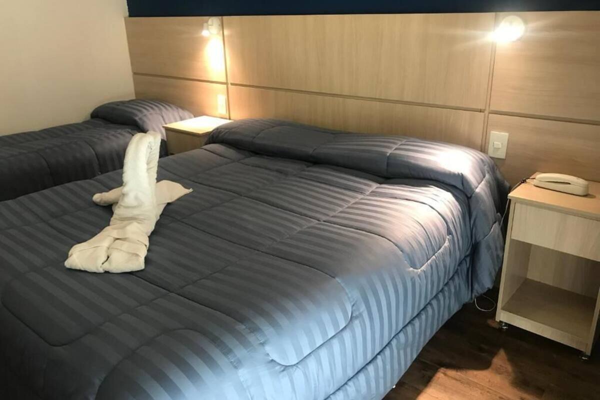 Habitación de hotel con dos camas, una mesita de noche con lámpara y un pequeño armario de madera.