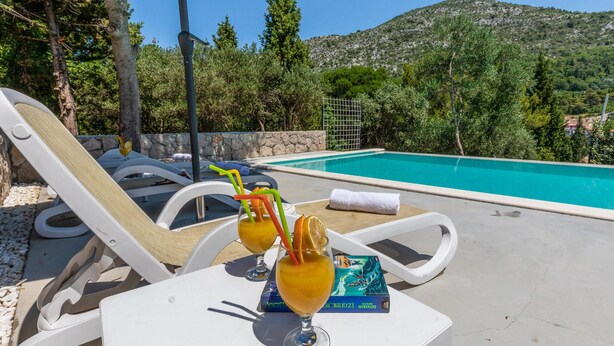 A poolside area with lounge chairs, a table with a drink and a book, and a clear view of a swimming pool and mountains in the background. - Gorgeous home in Kremena (Kremena)