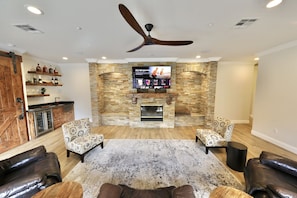 A living room with a stone fireplace, two patterned armchairs, a leather sofa, and a ceiling fan.
