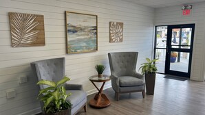 A lobby with a grey upholstered armchair, a round glass table with a plant, and a large painting on the wall. - The Ludlam (Sea Isle City)