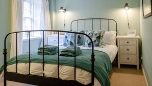 A bedroom with a metal bed frame, a white nightstand with drawers, a lamp, and a window with curtains. - Kirk Wynd Cottage - Classic Charm (Angus)