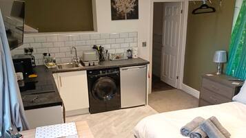 Habitación compacta con cama, una mesita, lavadora y kitchenette.
