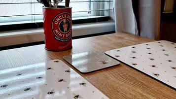 Una mesa con un mantel individual estampado con abejas, un recipiente con utensilios y una ventana con persianas.