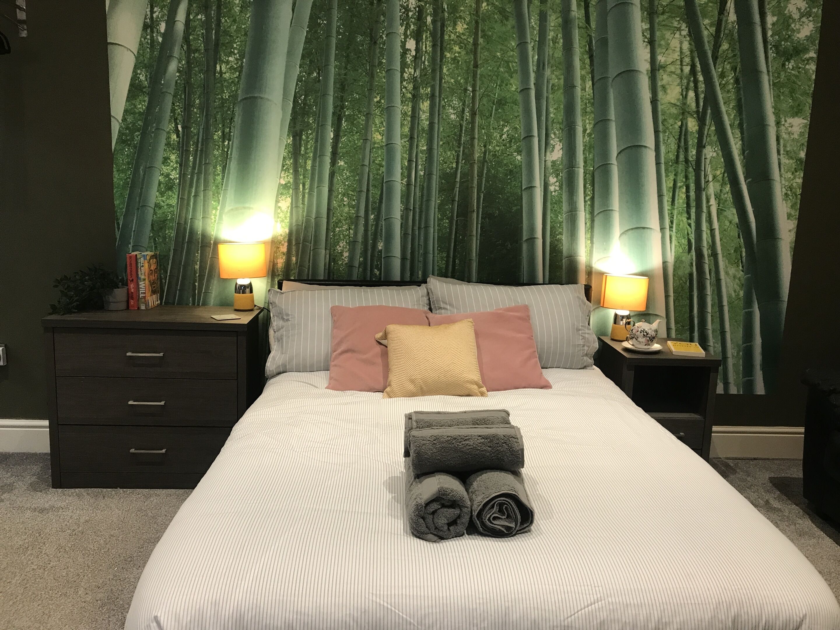 A bedroom with a large bed, two bedside tables with lamps, a dark wooden dresser, and a wall mural of bamboo forest.