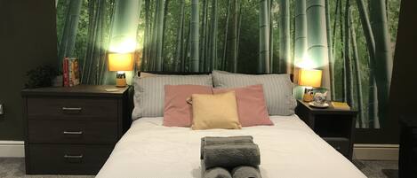 A bedroom with a large bed, two bedside tables with lamps, a dark wooden dresser, and a wall mural of bamboo forest.
