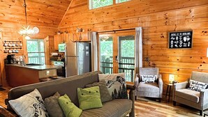 Ein Holzblock-Interieur mit einer Couch, Sesseln, einem Deckenventilator, einer Küchenzeile und einem großen Fenster.
