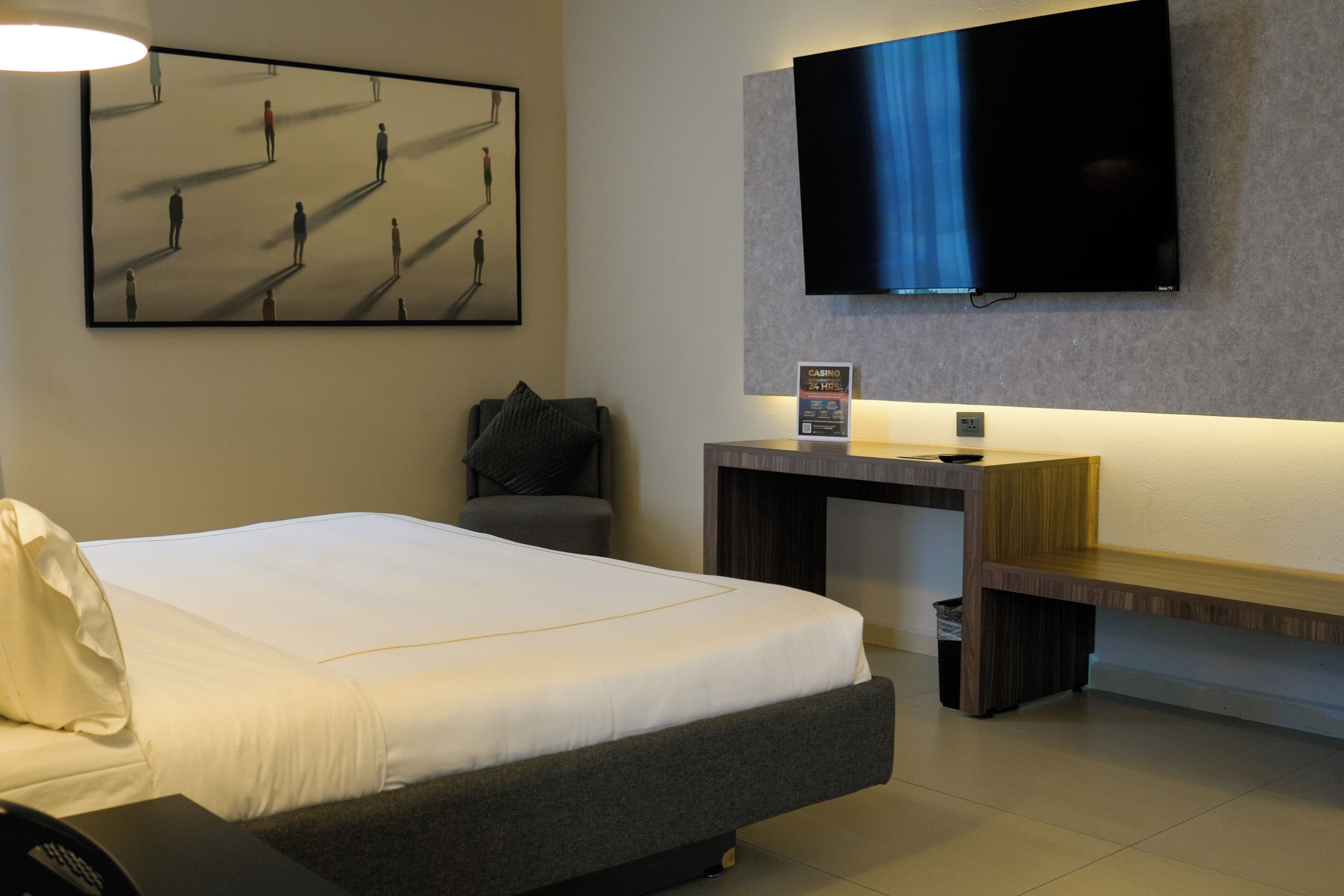 A hotel room with a bed, a desk, a TV mounted on the wall, and a painting on the wall.