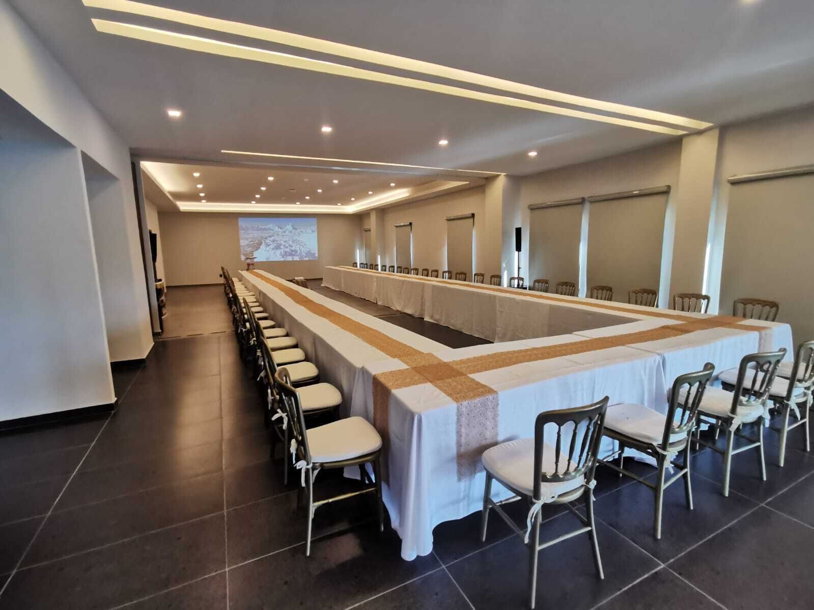 A conference room with a long table and chairs, equipped with a projector screen and ceiling lights.