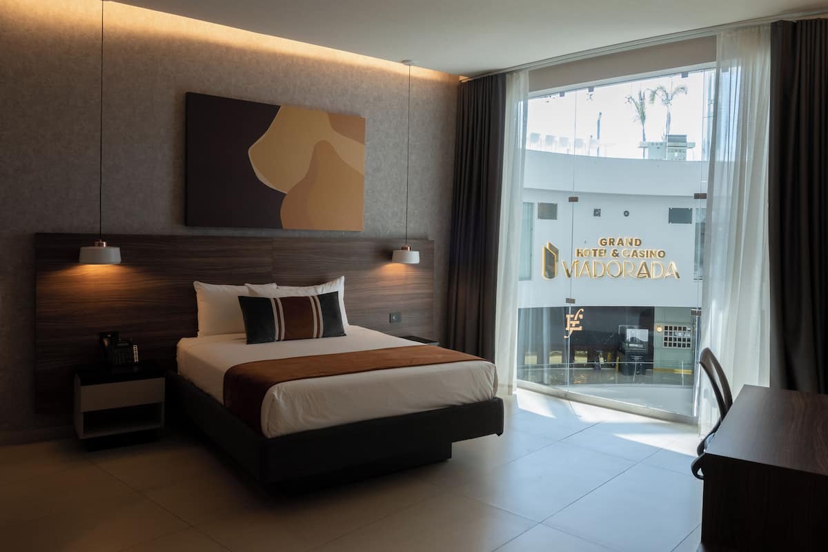 Una habitación de hotel moderna con una cama grande, lámparas de noche, vistas al Grand Hotel & Casino Vidorada y un área de comedor.