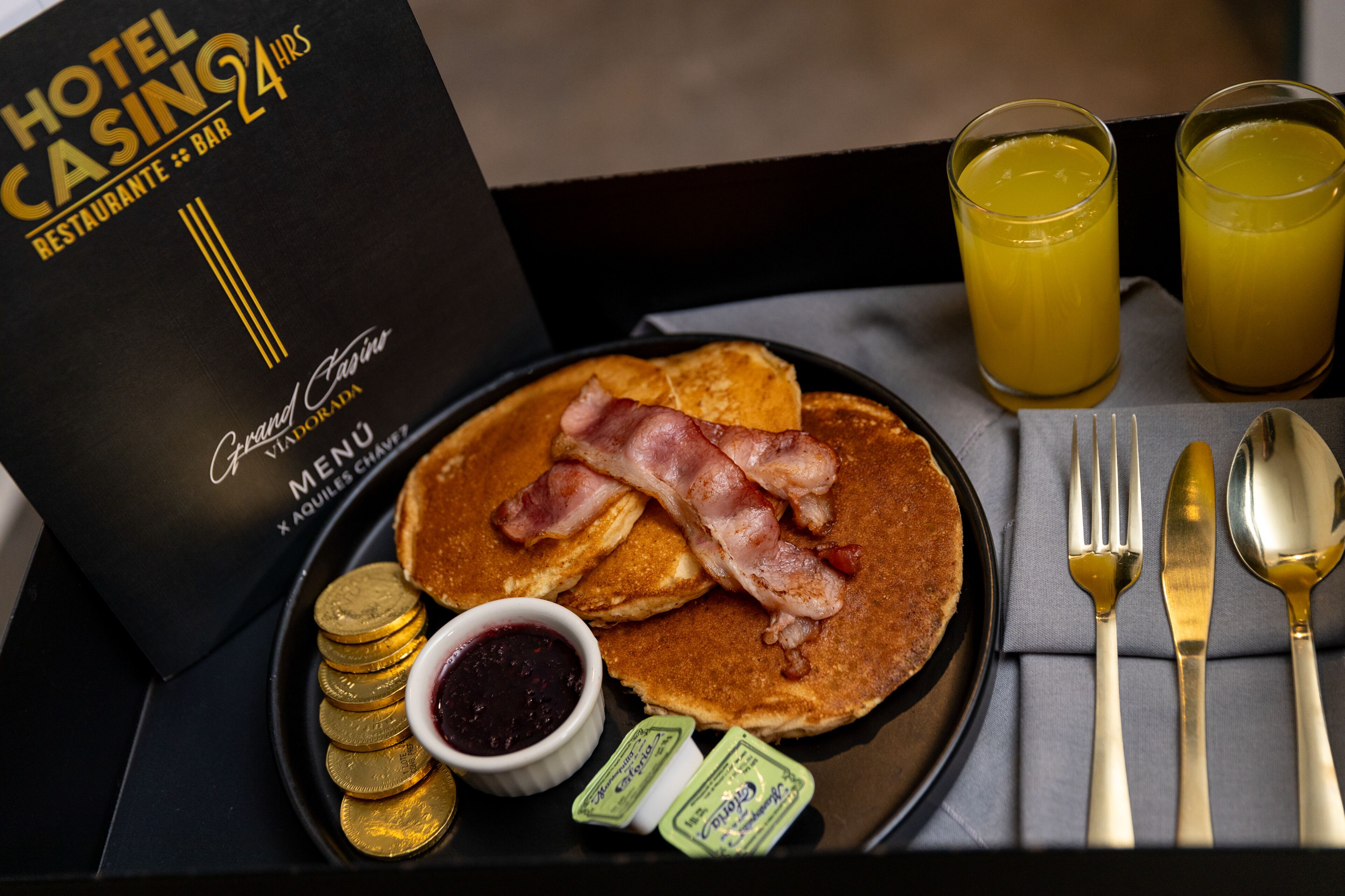 A breakfast tray with pancakes, bacon, gold coins, jam, butter packets, and a menu for a hotel casino restaurant.