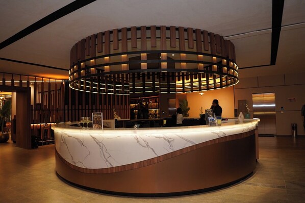 A modern reception desk with a marble top, surrounded by a wooden panel design and ambient lighting. - Hotel Casino Grand Vía Dorada (Pachuca)
