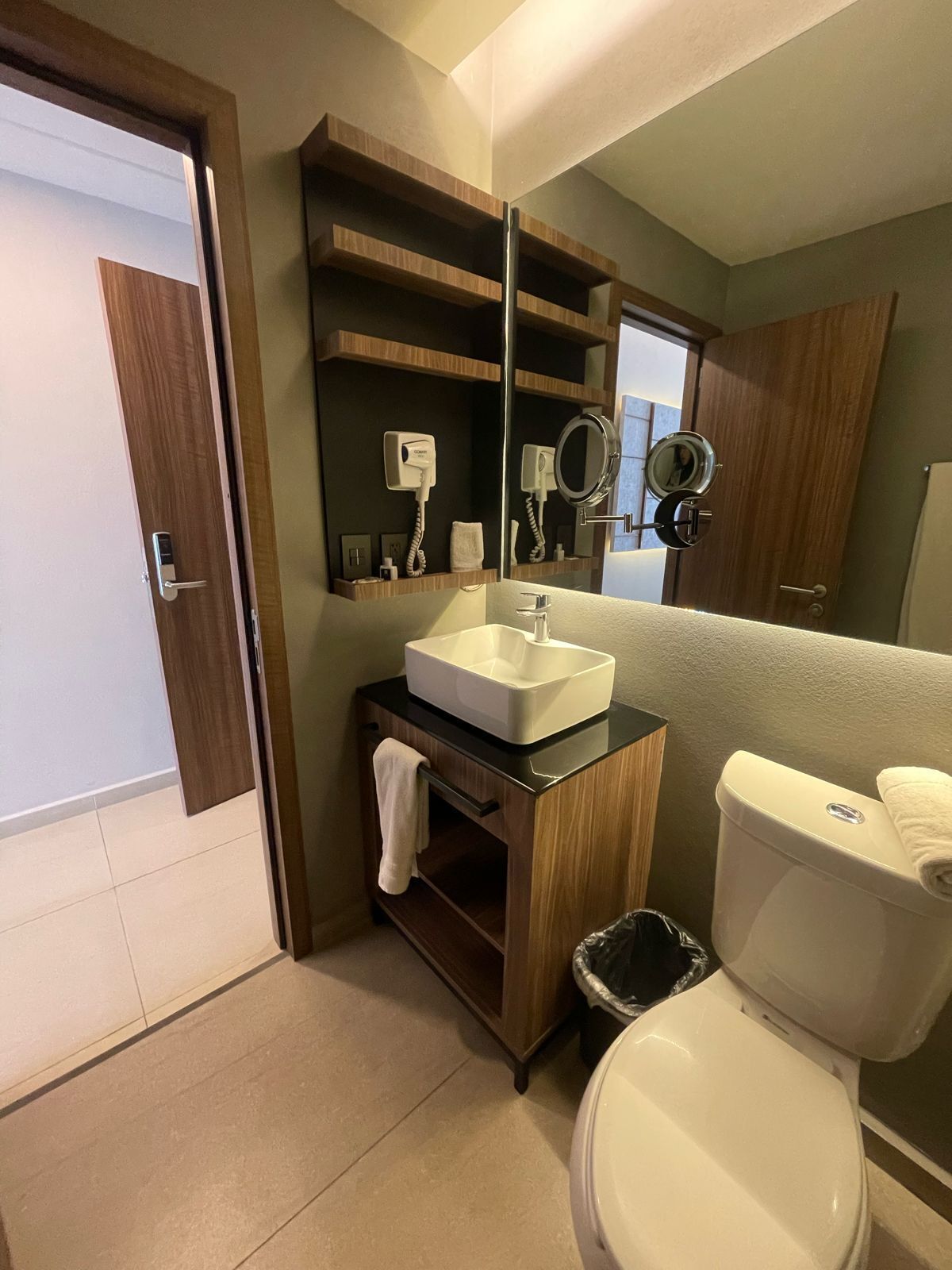 Une salle de bain moderne avec un lavabo blanc, des toilettes et un miroir au-dessus du lavabo. On trouve des étagères en bois au-dessus du lavabo et une poubelle à côté des toilettes.
