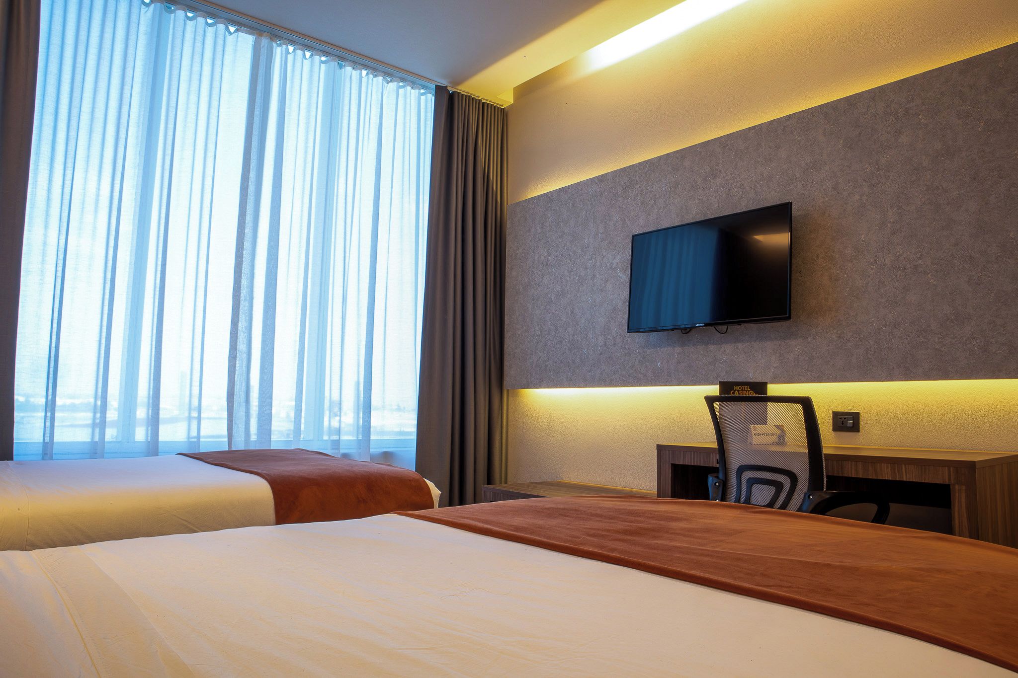 Habitación de hotel con cama, escritorio, silla, televisión y ventana con cortinas.
