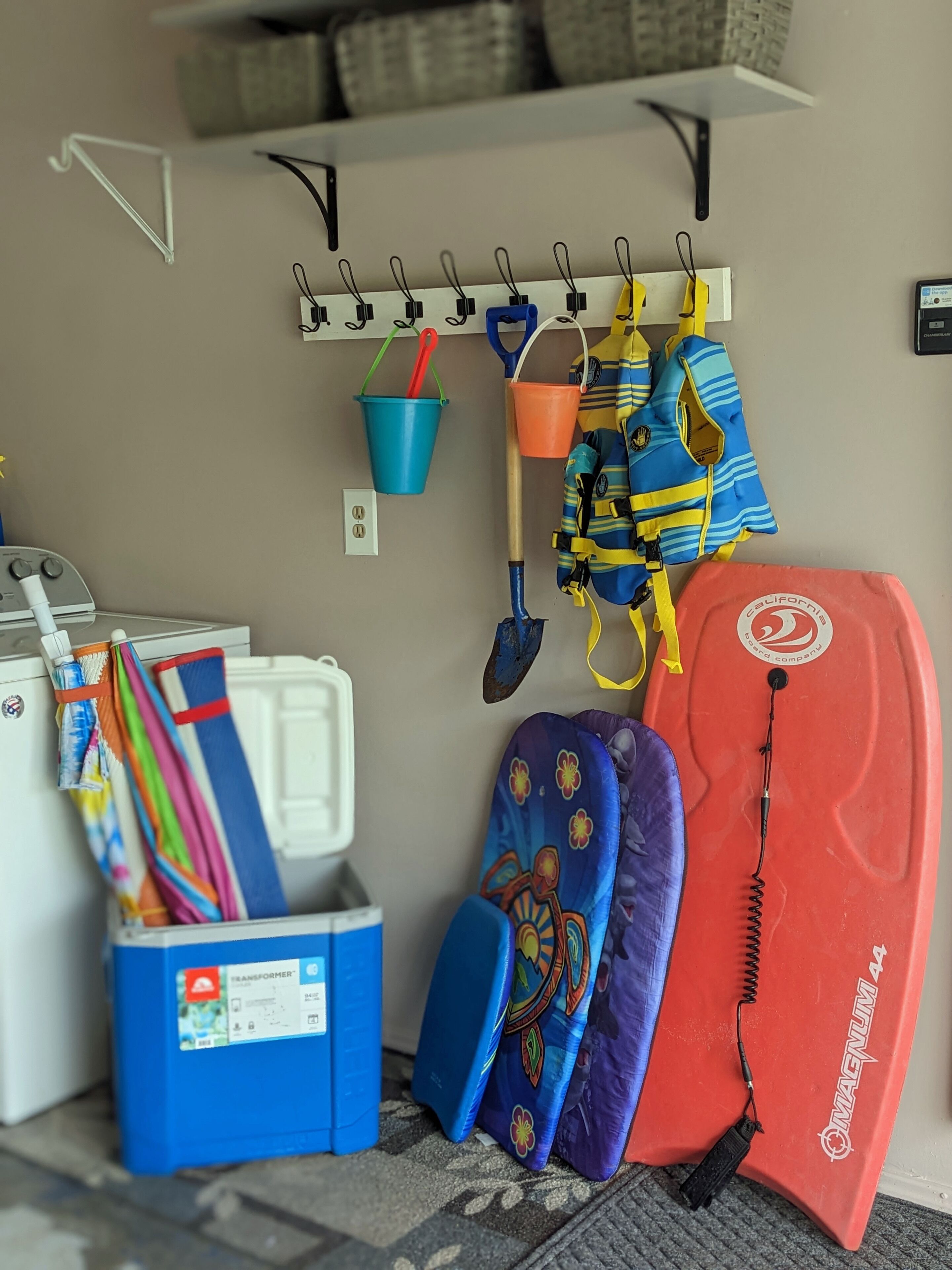 Un espace de rangement avec une étagère blanche sur laquelle sont accrochés divers objets, une glacière bleue et une planche de surf rouge.