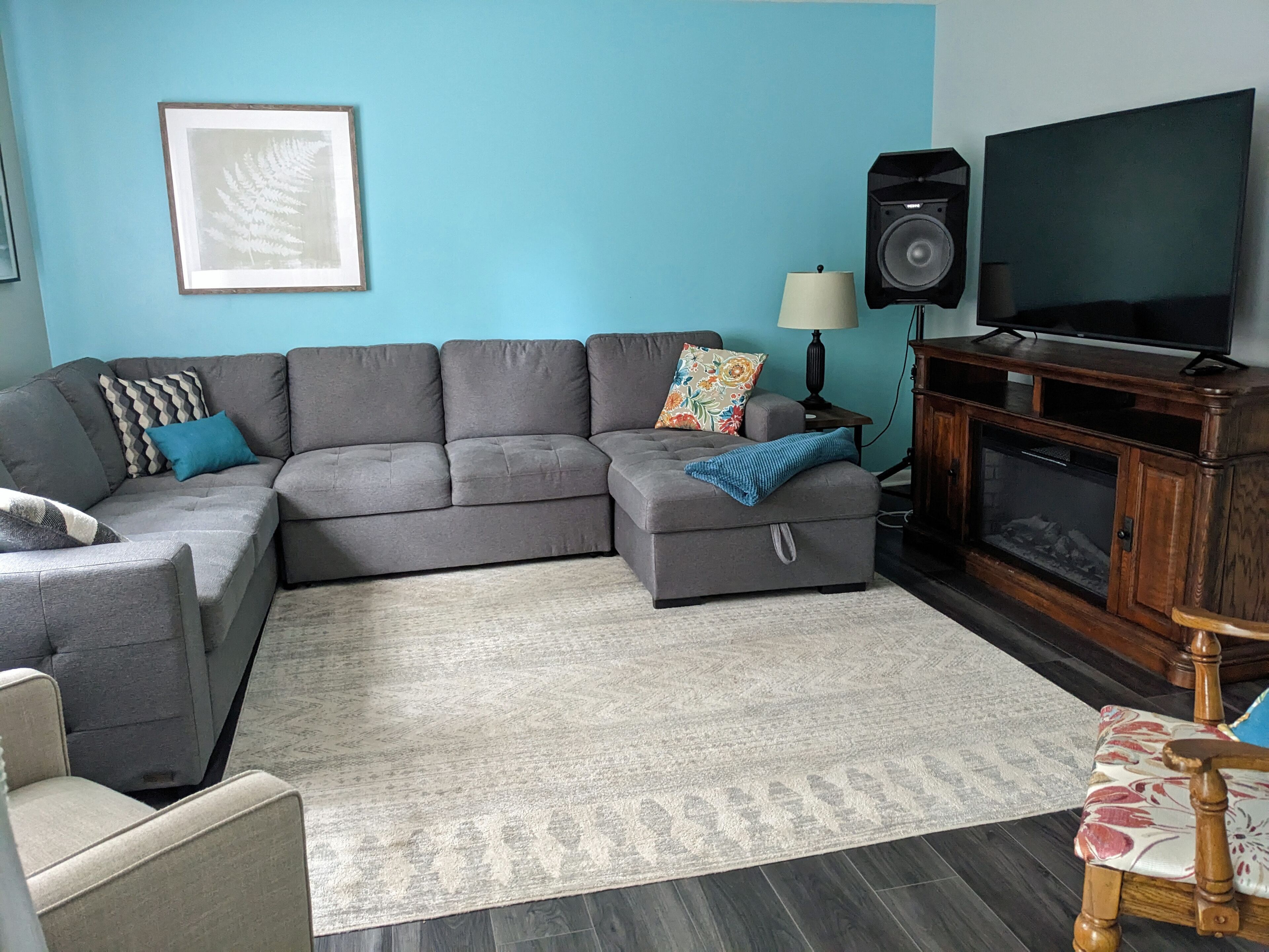 A living room with a grey sectional sofa, a small wooden cabinet with a television, a framed picture on the wall, and a patterned rug.