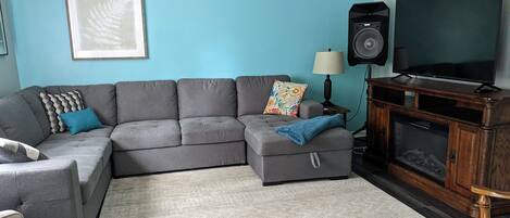 A living room with a grey sectional sofa, a small wooden cabinet with a television, a framed picture on the wall, and a patterned rug.