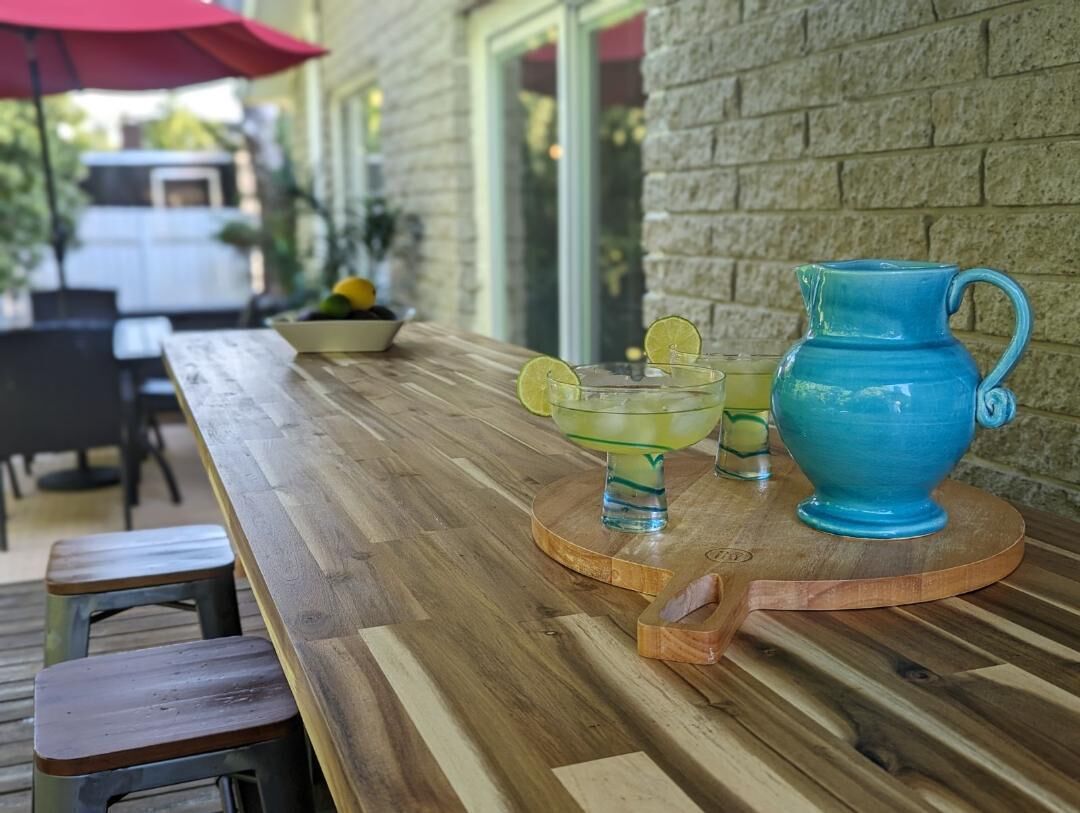 A wooden table with a blue pitcher and a glass pitcher, a bowl with fruits, and a red umbrella.