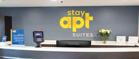 Ein Empfangstresen mit einem Schild 'stay apt SUITES', einem ViewSonic-Monitor, einem Zahlungsterminal und einer Vase mit Blumen.