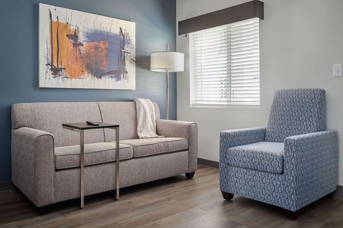 A living room with a grey sofa, a patterned armchair, a side table, a floor lamp, and a large abstract painting on the wall.
