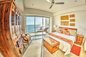 A spacious bedroom with a large bed, a wooden cabinet, a chair, and a view of the ocean through a large window. - Beachfront 2BR w Office & Panoramic Views: Nova105 (Bucerías)
