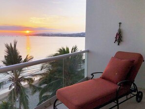 A balcony with a red chair, a railing, and a view of the sunset over water. - Beachfront 2BR w Office & Panoramic Views: Nova105 (Bucerías)