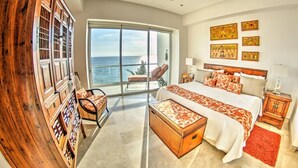 Un dormitorio espacioso con una cama grande, un armario de madera, una silla y vista al océano a través de un ventanal amplio.