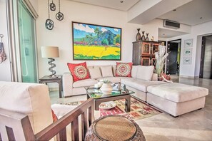 A spacious living room with a large sofa, a coffee table, and a painting on the wall. - Beachfront 2BR w Office & Panoramic Views: Nova105 (Bucerías)