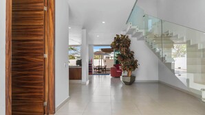 Un interior moderno con una puerta de madera, una escalera con barandilla de cristal, una planta en maceta y una zona de estar con mesa y sillas.