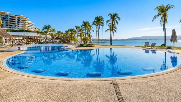 Una piscina amplia y transparente con sillas de descanso y palmeras, rodeada por un edificio de resort.